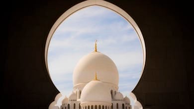 Captivating view of white domes of Sheikh Zayed Grand Mosque through an archway against the blue sky.